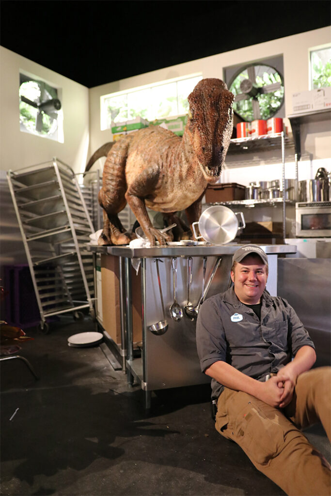 A man in a brown-ish-gray button up shirt with a white name tag reading "PHIL" over a black undershirt, brown pants, and a brown-ish-gray hat, smiles as he sits with his back against a kitchen counter in a recreation of a scene from "Jurassic Park." His fingers are interlocked, with his hands in his lap. Hanging from the front of the counter, to the man's right, are pieces of kitchenware like spoons and ladles. The kitchenware and counter are both silver. On top of the counter stands a large, brown dinosaur figure. It is surrounded by various silver bots and pans, laying scattered across the countertop. Behind the counter is a kitchen rack leaning over, and silver shelves with various kitchen supplies and containers or cans of food and ingredients.  Near the right of the photo is a silver microwave. Near the top of the visible walls are rectangular windows showing a sunny outside and green plants, and fans with windows to the outside too. The floor is black, and has a white plate seemingly thrown to the ground, and the ceiling is black.