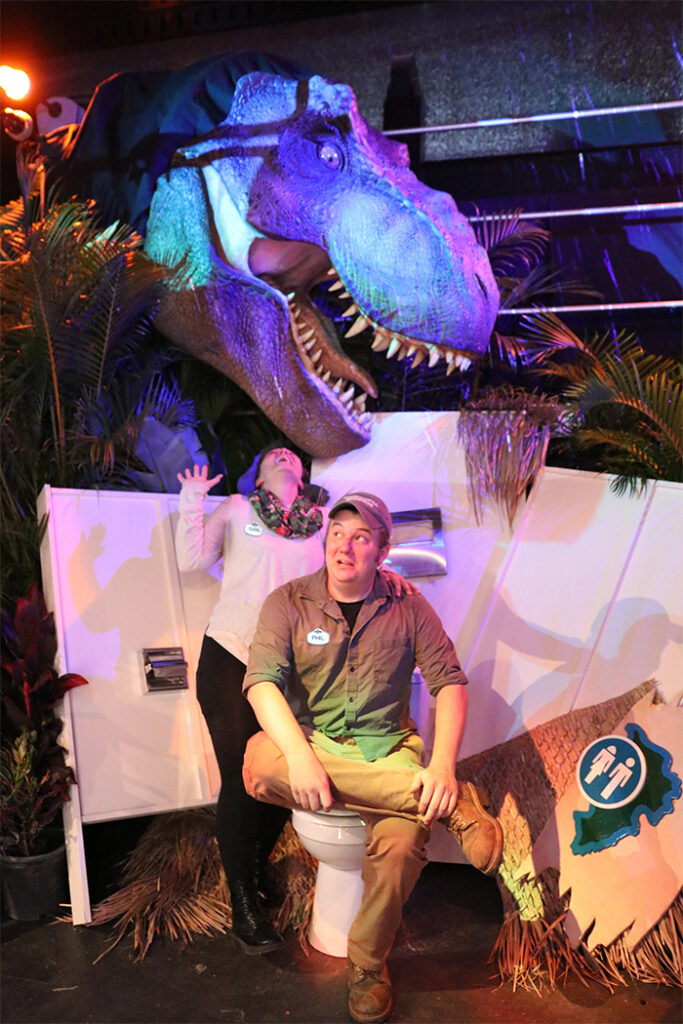 In between plants and a gated railing is the giant head of a dinosaur. It sits right above a set of a destroyed bathroom stall, with a closed toilet, that a man sits on and a woman stands behind. The man is wearing a brown-ish-gray shirt with a black undershirt, a gray hat, and has a white name tag reading "Phil" pinned to his chest. The woman is wearing a pink shirt, a flowery scarf, and has a white name tag reading "Sara." The woman has her left hand on the man's shoulders, and her right arm bent at the elbow, with her palm facing up. She looks up, with her mouth open. The man looks to his right. Both pretend to be scared. The pair is surrounded by broken walls with a toilet paper dispenser, a seat cover dispenser, and a bathroom sign with the silhouette of a man and a woman.