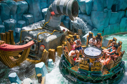 A view of a full raft floating on Popeye & Bluto's Bilge-Rat Barges. Surrounding the raft, made to look like it was made of wood, which has netting between each pair of seats, a plastic circular center, and turquoise linings, are crashing waves. Behind a gate, made to look like logs of wood, is a pipe, spouting water onto the guests in the raft. The front of a boat can be seen on the far left of the photo. A wall made of blue rocks stands in the background. It appears to be sunny outside, where the photo was taken.