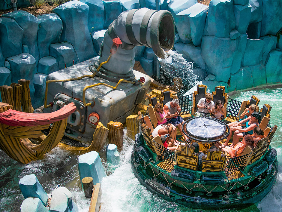 A view of a full raft floating on Popeye & Bluto's Bilge-Rat Barges. Surrounding the raft, made to look like it was made of wood, which has netting between each pair of seats, a plastic circular center, and turquoise linings, are crashing waves. Behind a gate, made to look like logs of wood, is a pipe, spouting water onto the guests in the raft. The front of a boat can be seen on the far left of the photo. A wall made of blue rocks stands in the background. It appears to be sunny outside, where the photo was taken.