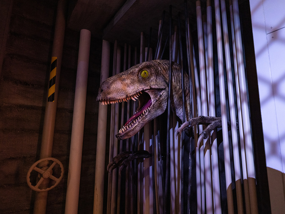A close-up of a giant raptor figure, with a visible yellow eye, an open mouth full of sharp teeth and a long tongue, and sharp claws, popping out of what looks like a gated cell. Behind the figure are pipes, one of which with a steering wheel and a yellow-and-black striped sticker.