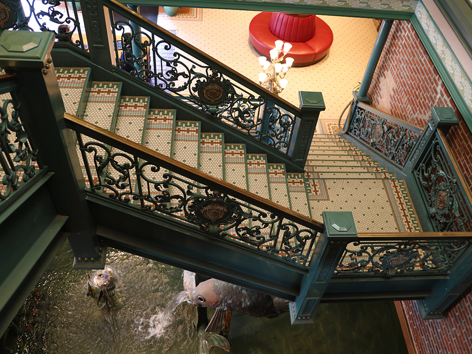 A view from above of a tiled, zig-zag staircase in Lombard's Seafood Grille. The green-and-gold railings have an ornate, metal design, with a green-and-gold pendant near the center of each four-sided section. Green dots scatter the steps, along with a tiled pattern. The battern continues on the ground floor after the staircase ends, and a red circular couch sits at the center of that floor. On one corner of the staircase is a multi-lighted lamp. Under the staircase is a fountain, with water flowing from a fish, appearing to be made of metal or stone. The walls are seemingly made of red bricks.