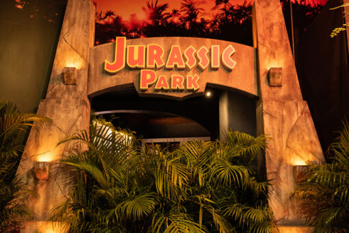A view of an archway in the Jurassic Park Tribute Store at Universal Orlando Resort. The arch itself is seemingly made of a brown rock material, and four lanterns sit on the archway, two on the left and two on the right. The text "Jurassic Park" is blocky and orange, with a yellow border. Under the archway sit plants, with leaves extending forward beyond the archway's front. Above the arch is a dimly lit, red-and-orange-hued background, with the silhouettes of trees and bushes.