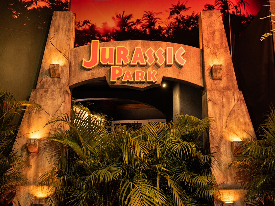 A view of an archway in the Jurassic Park Tribute Store at Universal Orlando Resort. The arch itself is seemingly made of a brown rock material, and four lanterns sit on the archway, two on the left and two on the right. The text "Jurassic Park" is blocky and orange, with a yellow border. Under the archway sit plants, with leaves extending forward beyond the archway's front. Above the arch is a dimly lit, red-and-orange-hued background, with the silhouettes of trees and bushes.