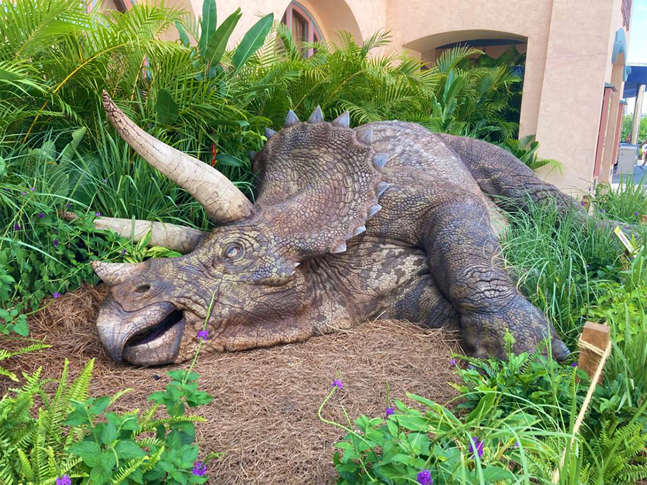 A view of the triceratops figure from outside the "Jurassic Park" Tribute Store at Universal Studios Florida laying down in a patch of dirt, hay, and green plants. Behind the patch are beige buildings and the hints of a sunny sky.