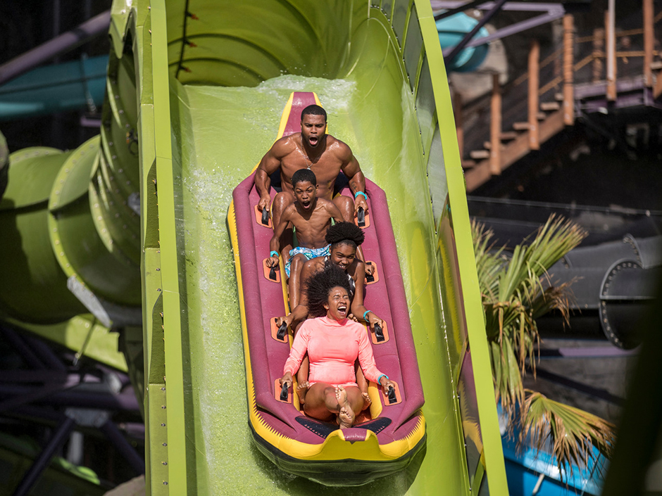 A group of four inside of a purple raft with yellow borders, going down a green water slide. From front to back in the raft: a woman in a pink, long-sleeve top, with her mouth open mid-scream, not wearing shoes; a woman with clothes blocked by the woman in front, leaning forward; a boy wearing purple swim trunks, screaming; and a man with clothes blocked by the boy in front, with his mouth open. The slide begins as a tube, before opening up to a standard slide structure. To its left is a palm tree, and in the background are various tubes, and staircases.
