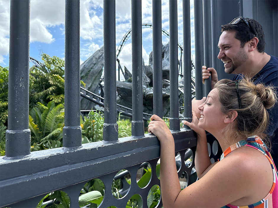 A woman with blonde hair and sunglasses on her head wearing a multi-colored outfit and a man with dark hair in a navy shirt with sunglasses on his head smile while holding onto a blue-ish-gray gate outside of Jurassic World VelociCoaster. In front of the coaster, which has spiky rocks surrounding it, are various green plants,