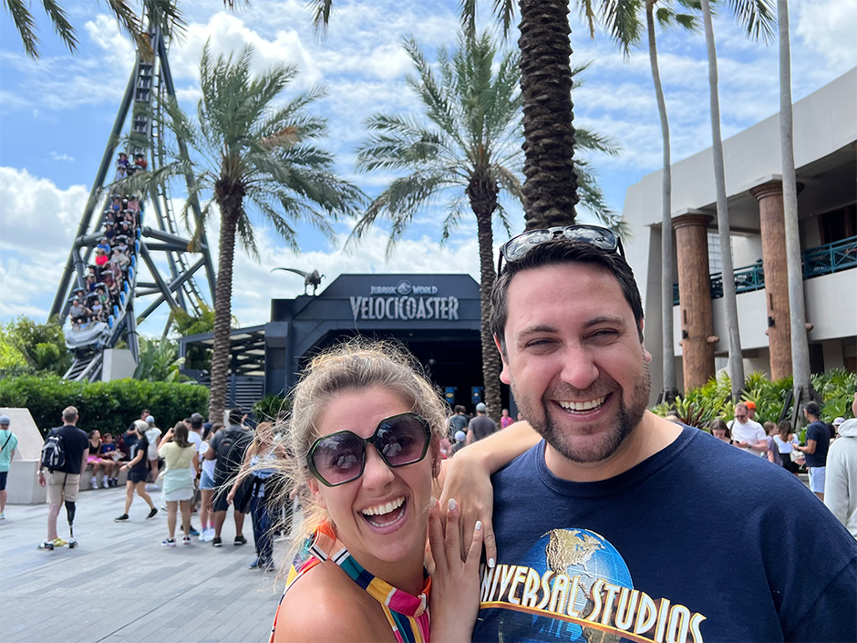 A woman with blonde hair wearing sunglasses and a multi-colored dress puts her arms on the right shoulder of a man with dark hair in a navy Universal Studios shirt, with the globe logo, and sunglasses on his head. The woman's fingers are interlocked with each other, and the two people are smiling. They stand outside the entrance to Jurassic World VelociCoaster at Universal Islands of Adventure. A lift hill for the roller coaster can be seen to the left of the entrance, with people sitting in the vehicle. A crowd walks behind the pair. There are a few seemingly palm trees in the background, as well as a blue sky with several white clouds. To the right of the pair is seemingly the side of a mostly off-white building, with brown columns.