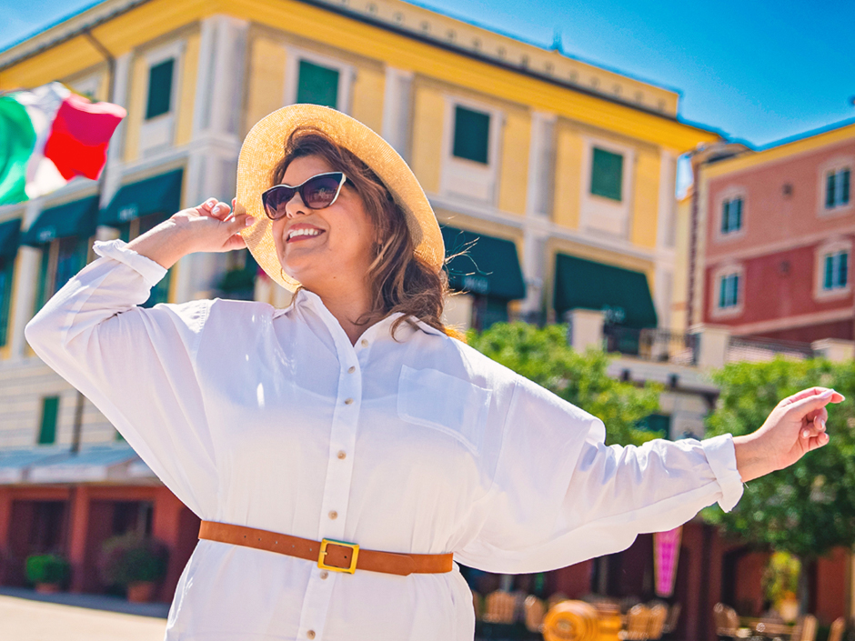 Woman with brown hair, wearing a hat, sunglasses, and a white button-up shirt, standing in front of Italian-inspired architecture at Loews Portofino Bay Hotel with a smile on her face