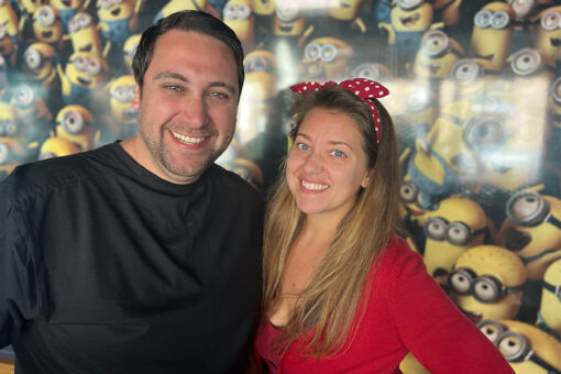 Two people stand in front of a wall of yellow Minions, which are wearing goggles and overalls. On the right is a man in a black shirt, and on his left is a woman in a red, long-sleeve shirt with a red headband tied in a bow. They both smile, and seem to have one arm wrapped around each other.