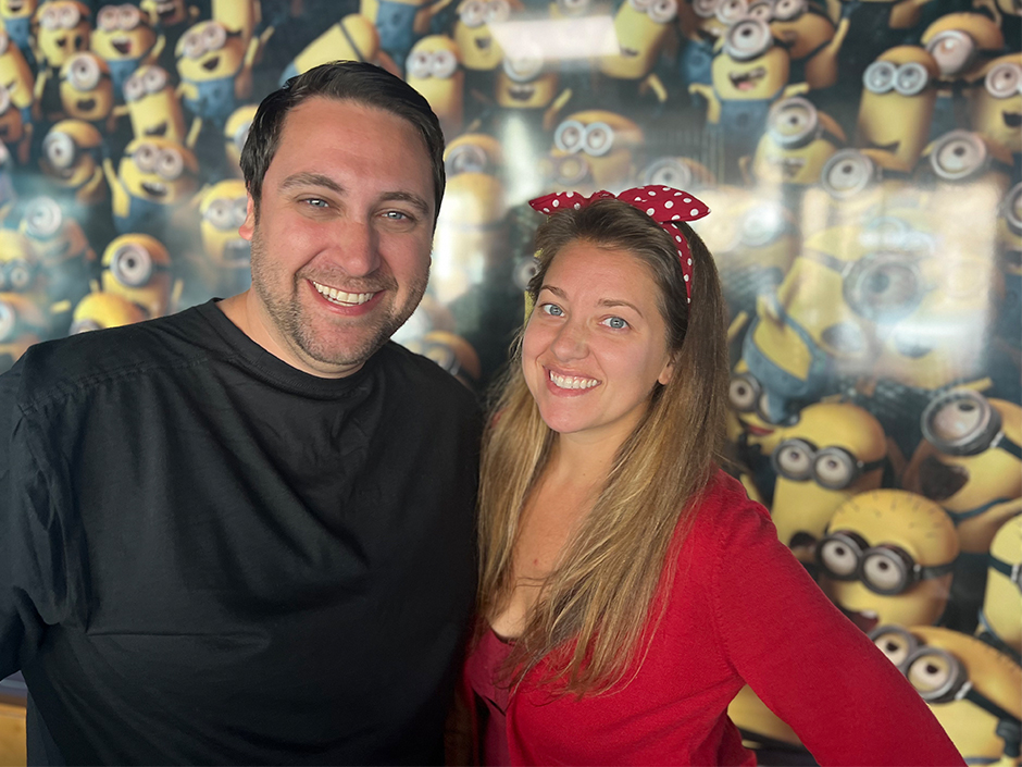 Two people stand in front of a wall of yellow Minions, which are wearing goggles and overalls. On the right is a man in a black shirt, and on his left is a woman in a red, long-sleeve shirt with a red headband tied in a bow. They both smile, and seem to have one arm wrapped around each other.