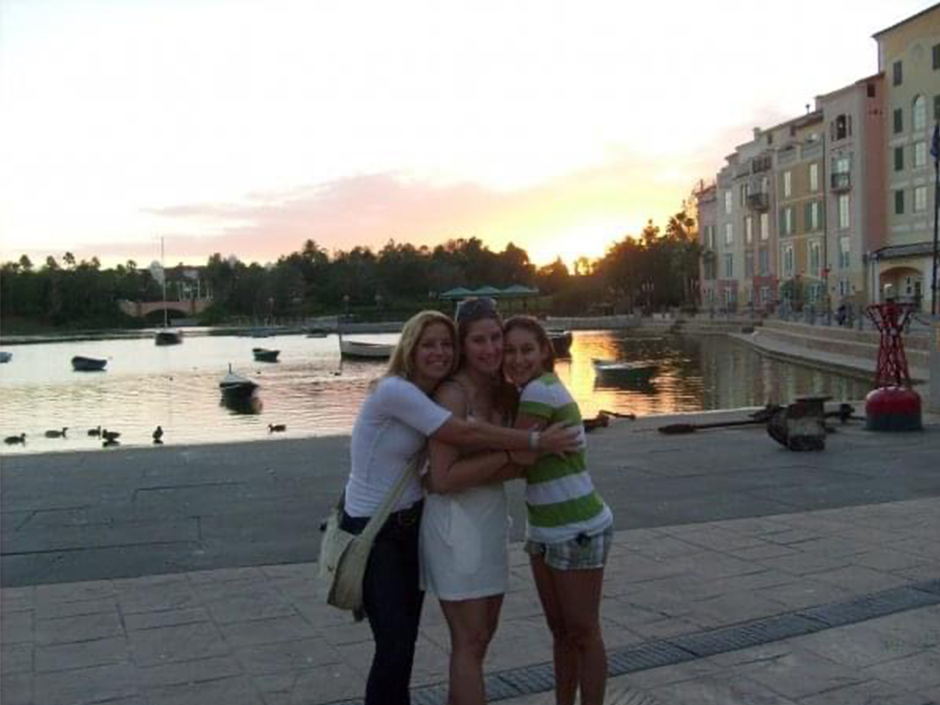 Three women standing in front of a sunset over the water at Loews Portofino Bay Hotel, hugging and smiling
