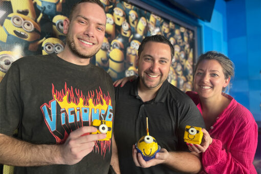 Three people stand in front of a wall of yellow Minions, which are wearing goggles and overalls, with a blue, rectangular pattern to its left. On the right is a man in a black T-shirt that says "Vicious 6" in blue on top of a fire background. In the center is a man in a black button-up shirt. On the left is a woman in a pink, long-sleeve shirt. They each hold a Minion-themed dessert in front of them, the outer two being cube-shaped and the center one being spherical, with a stick popping out the top.