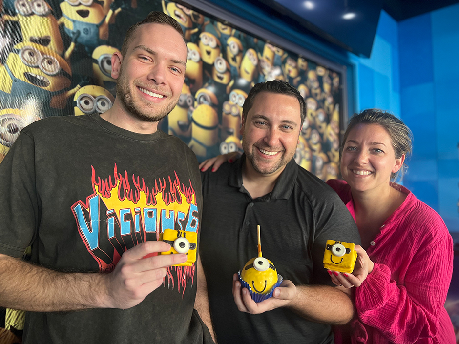 Three people stand in front of a wall of yellow Minions, which are wearing goggles and overalls, with a blue, rectangular pattern to its left. On the right is a man in a black T-shirt that says "Vicious 6" in blue on top of a fire background. In the center is a man in a black button-up shirt. On the left is a woman in a pink, long-sleeve shirt. They each hold a Minion-themed dessert in front of them, the outer two being cube-shaped and the center one being spherical, with a stick popping out the top.