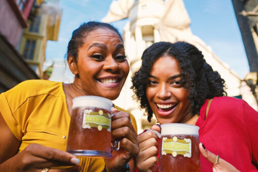 Two women smile while holding glass mugs of butterbeer, a brown-ish drink, in Diagon Alley at Universal Studios Florida. The woman on the left wears a yellow shirt, rings on both hands, a watch on her left wrist, and blue polish on the tops of her fingernails. The woman on the right wears a red shirt, one visible ring on her left hand, and black nail polish. The glass mugs have a green-ish-brown logo on them, with off-white decoration on the border of the wavy square shape, and a wavy, green banner across the vertical center. On the green banner is the word "Butterbeer" in gold. There are multi-colored buildings to the woman in yellow's right, and behind the pair is the tall, marble-like Gringotts Bank. The wings of the marble-like dragon that sits on top of the building are also visible. The sky is blue with some faint clouds.