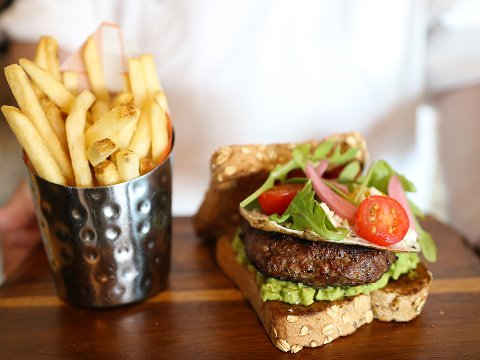 A metal cup with circular dents filled with french fries sits on a brown board-like plate, next to a burger. Instead of hamburger buns, there are two pieces of toast, with seeds scattered along the crusts. The top piece of toast leans against the back of the burger, which sits on top of the piece of toast laying flat on the board. There is avocado spread under the burger, and lettuce, tomato, onion, and other vegetables on top of it. The tray is being held by a blurred person in a white shirt.