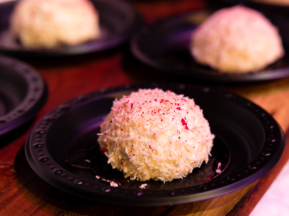 A white coconut snowball, lightly splattered in a red liquid, sits on a circular, black plate. Three other blurred, but identical, snowballs on plates sit near it on a wooden tabletop.