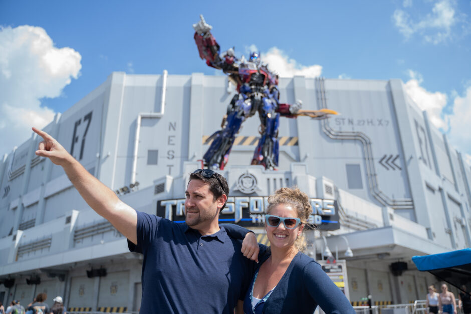 A man and a woman stand posing in front of Transformers The Ride - 3-D. The woman, wearing a dark blue overshirt over a white shirt and blue sunglasses, rests her right arm on the man's shoulder. The The man, wearing a dark blue polo, points to his right. He is matching the pose of the blue-and-red robot figure standing on the lower of the silver building's two roofs. The sky is blue with a couple clouds.