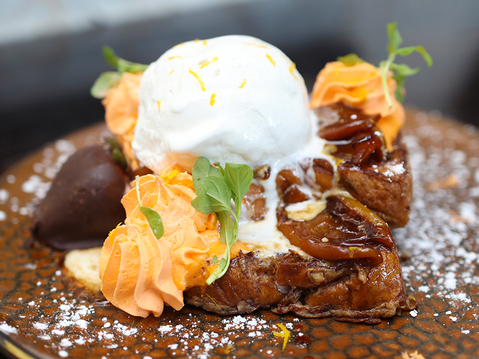 A close-up of a slice of brown, glazed french toast, topped with peaches, orange icing, green garnishes, and vanilla ice cream with orange shavings. Next to the french toast sits a chocolate-covered strawberry. The french toast and strawberry sit on top of a plate patterned with brown, rounded shapes, which is covered in powdered sugar. The background is blurred, and mostly dark gray with some white.