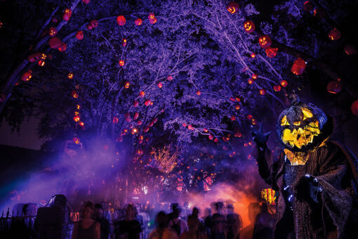 Crowd of people walking through outdoor scarezone at night, with puple light reflected on fog, red lights on trees and creepy scareactor dressed as a pumpkin on the right