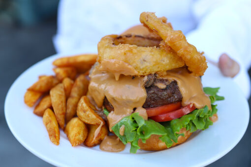 An arm in a white sleeve holding up a white plate. On the plate, there is a pile of potato wedges, on the left side of a burger sitting on a bottom bun. Under the burger is lettuce and tomato, and above it is a different meat, a melted cheese sauce, and two onion rings. The background is blurred and mostly brown.