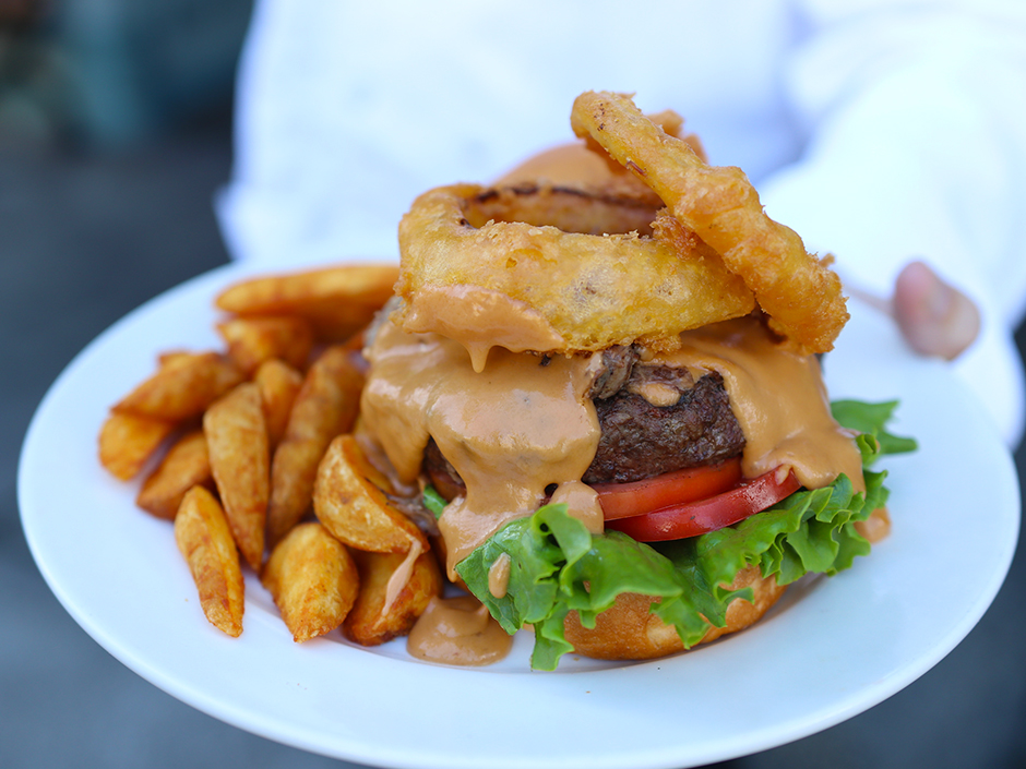 An arm in a white sleeve holding up a white plate. On the plate, there is a pile of potato wedges, on the left side of a burger sitting on a bottom bun. Under the burger is lettuce and tomato, and above it is a different meat, a melted cheese sauce, and two onion rings. The background is blurred and mostly brown.