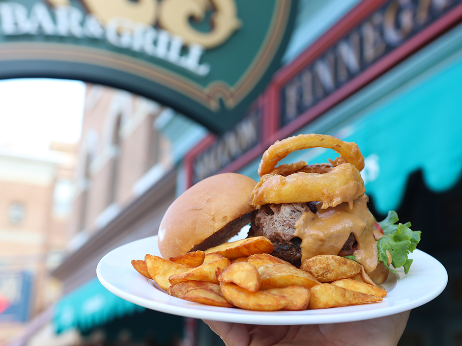 A hand holding up a white plate of food in front of Finnegan's Bar and Grill. On the plate, there is a pile of potato wedges, in front of a burger sitting on a bottom bun, with a top bun leaning onto it. Under the burger is lettuce and tomato, and above it is a different meat, a melted cheese sauce, and two onion rings. The building behind the plate, to its left, is blurred, with a turquoise awning, and a red-bordered, blue sign above the awning with brown-ish lettering. There is another sign in front of the awning, directly behind the plate, which is a darker green with a gold and red border. There are other blurry brown buildings behind the first, and the sky is sunny.