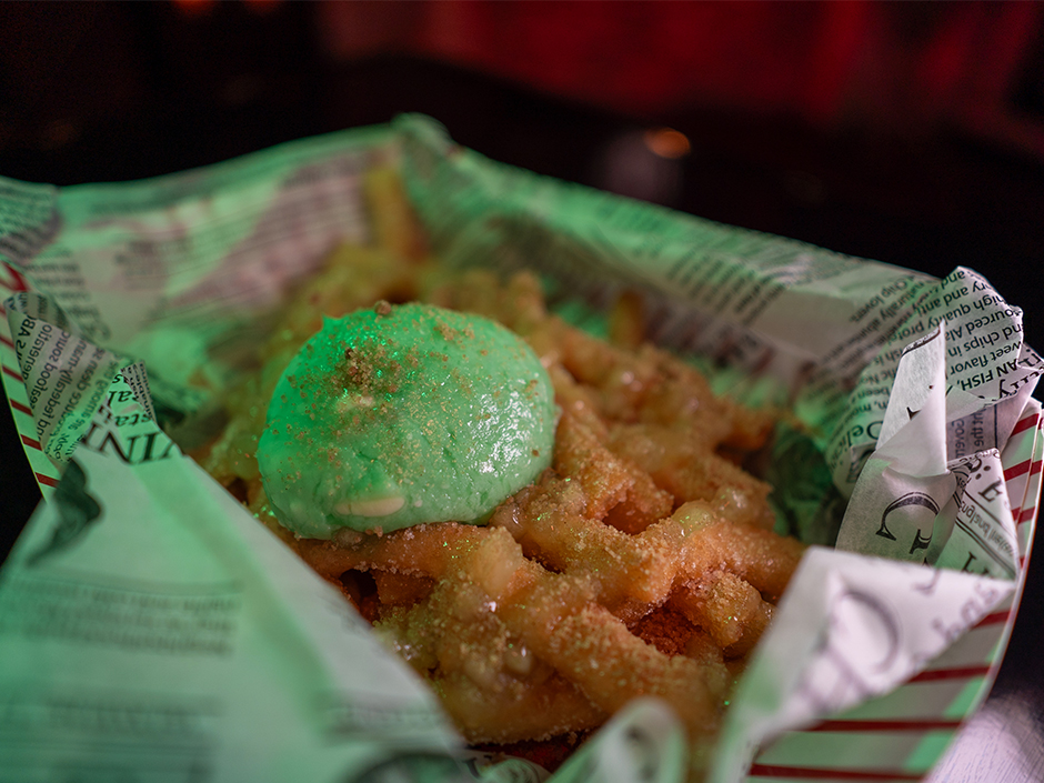 A basket, covered in newspaper, of fries, covered in seemingly cinnamon and sugar, and topped with green ice cream, which is also covered in seemingly cinnamon and sugar. The mostly red-and-black background is blurred.