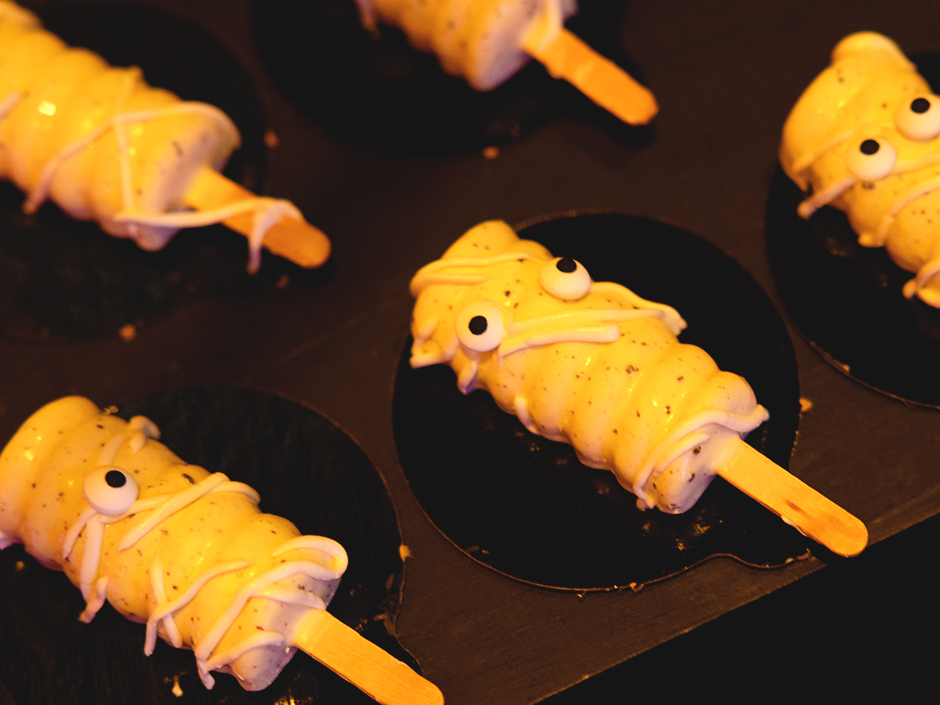 Five white, four-sided, round cake pops, with candy eyes and a white drizzle, skewered with a popsicle stick each and covered in cookie specks, each sit on a rounded, black platform. The eyes are only visible in the three in the front row of the five. and the one on the far left in front only has one eye. All five platforms sit on a dark brown, wooden shelf. 