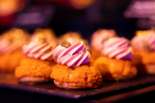 Several pumpkin-shaped puff pastries, topped with a seemingly pink frosting and green seeds, sit on a black tabletop. The background, along with the last row of pasties, is blurred.