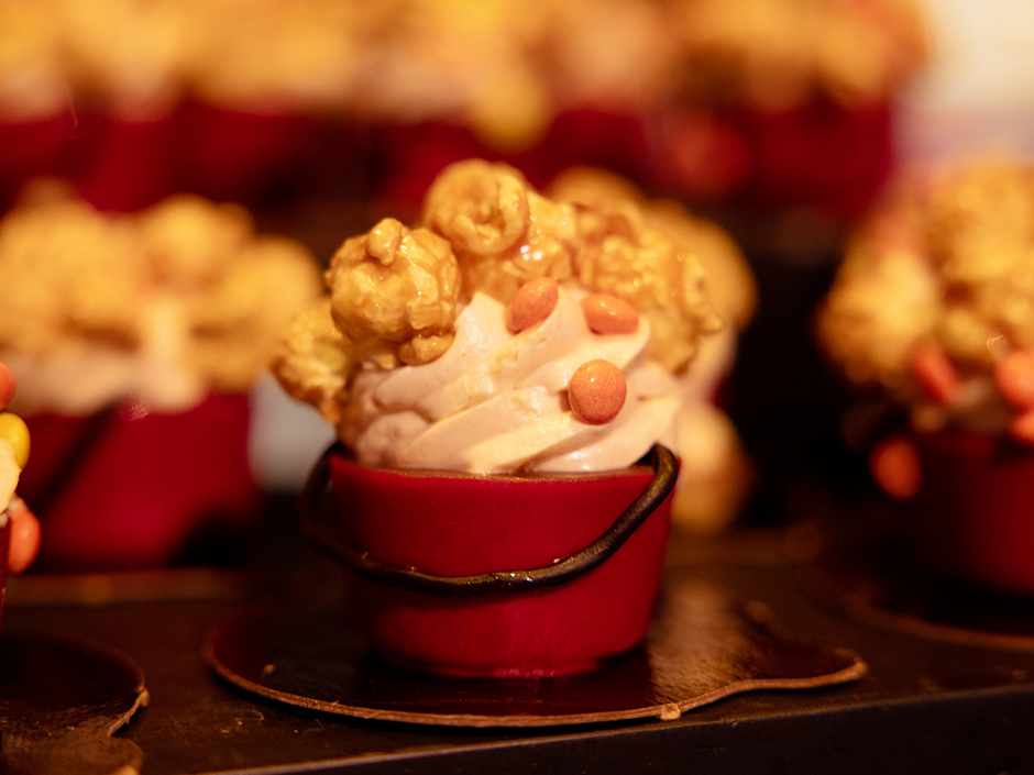 A red cup-like dessert, wrapped in a chocolate string, filled with an off-white cream, yellow circular candy, and caramel popcorn. It sits on a round, flat, black platform, which sits on a black shelf. Several matching blurred desserts surround it, and the rest of the background is blurred and mostly orange.
