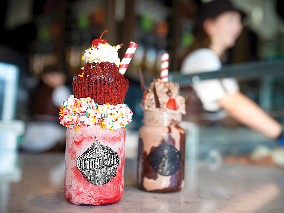 A red and white milkshake in a jar reading "Toothsome Chocolate Emporium," topped with whipped cream, sprinkles, and a red velvet cupcake.