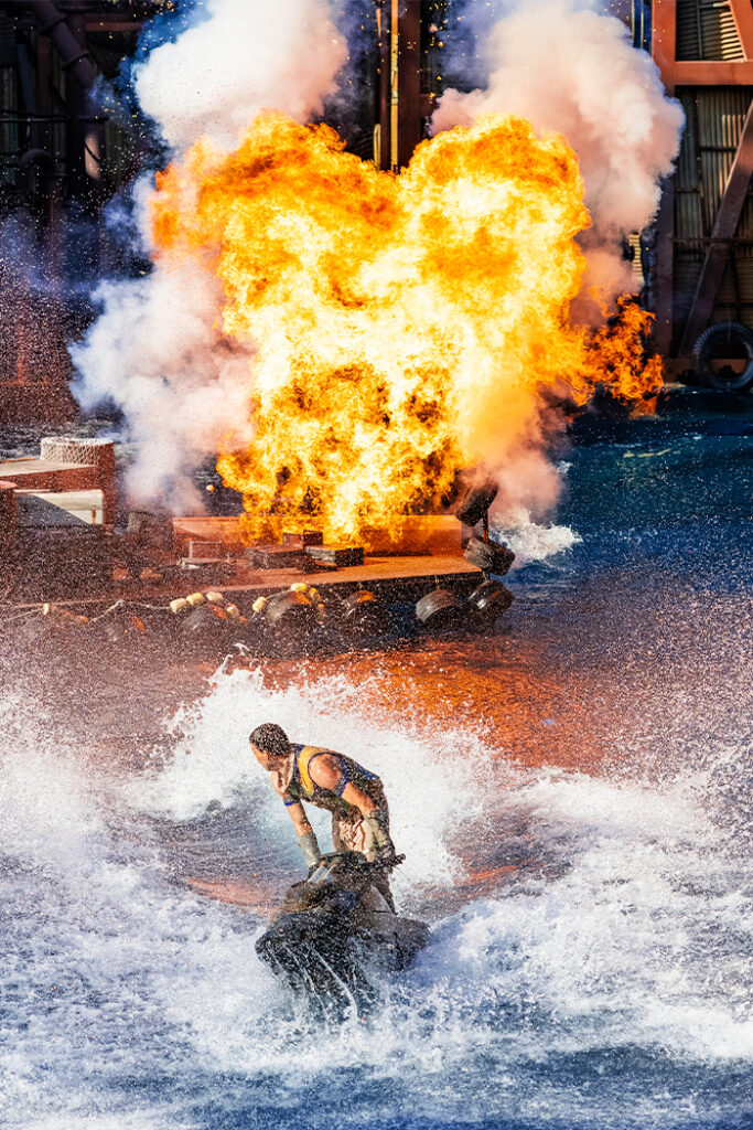 Man stands on jet ski looking back on large explosion behind him.