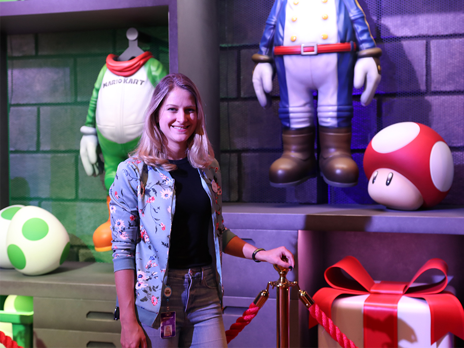 A blonde woman stands and smiles in the Mario Kart queue. 
