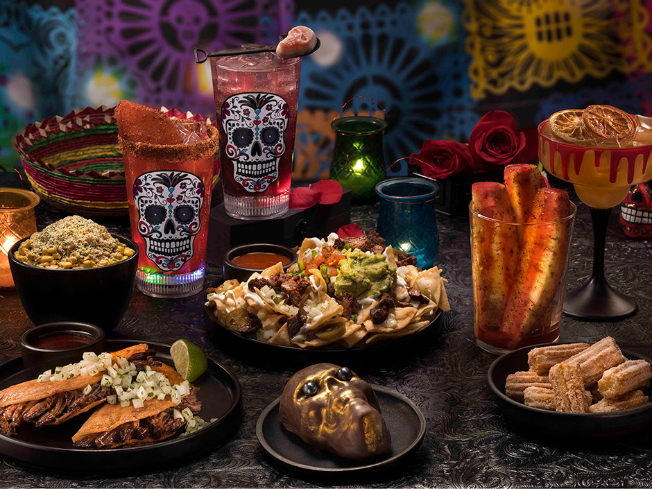 A spread of Dia De Los Muertos-themed food and drink, including tacos, nachos, and churros.