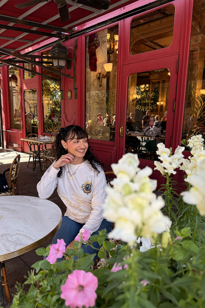 A guest smiles for a picture while sitting outside at Cafe L'air de la Sirene in The Wizarding World of Harry Potter - Ministry of Magic in Epic Universe