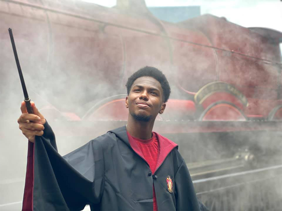 Man pointing a wand and wearing a Gryffindor robe posing in front of the Hogwarts Express.