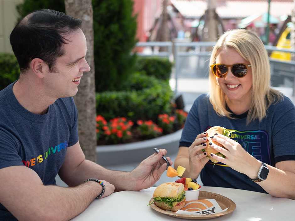 A man and woman wearing Love Is Universal shirts eating a Pride sandwich