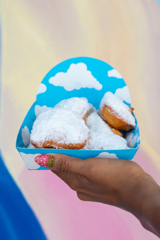 A hand holding up a plate of beignets in front of a rainbow-colored wall