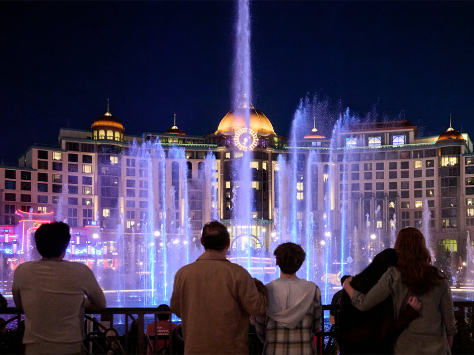 Guest watch the Epic Universe fountain show at night in front of Helios Grand Hotel