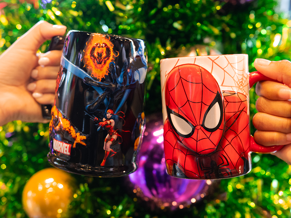 Two hands hold up mugs featuring Spider-Man and a variety of Marvel heroes in front of a Christmas tree