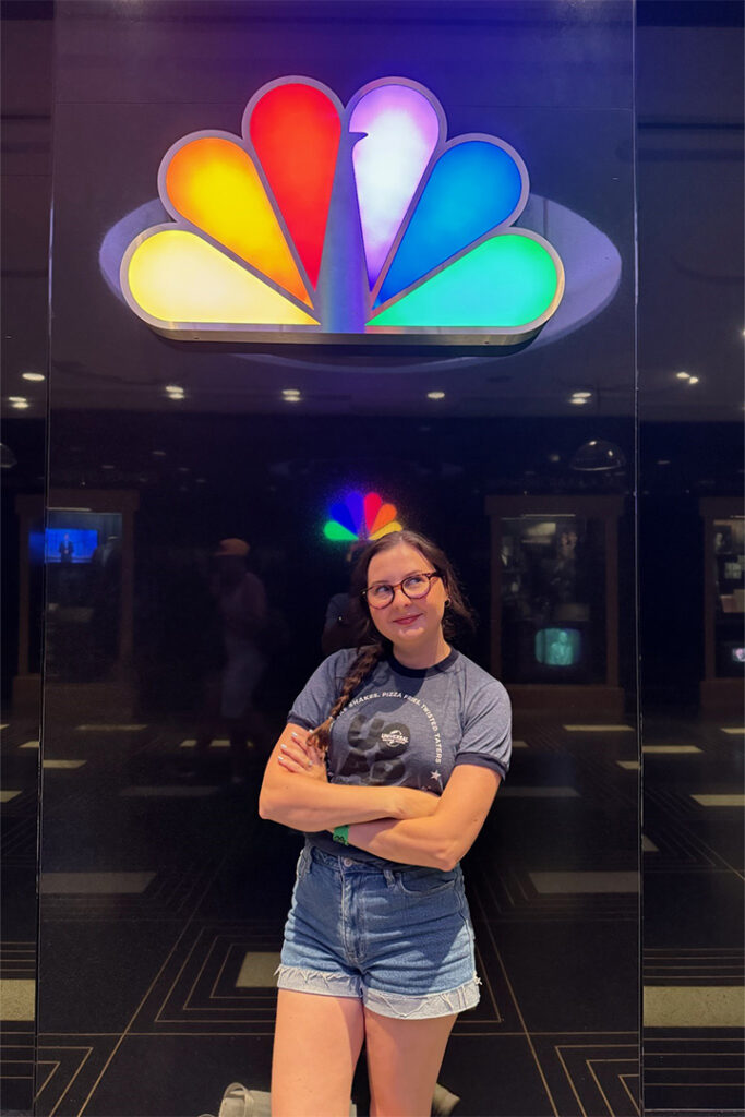 A guest smiles and poses under the NBC logo in the Race Through New York with Jimmy Fallon queue