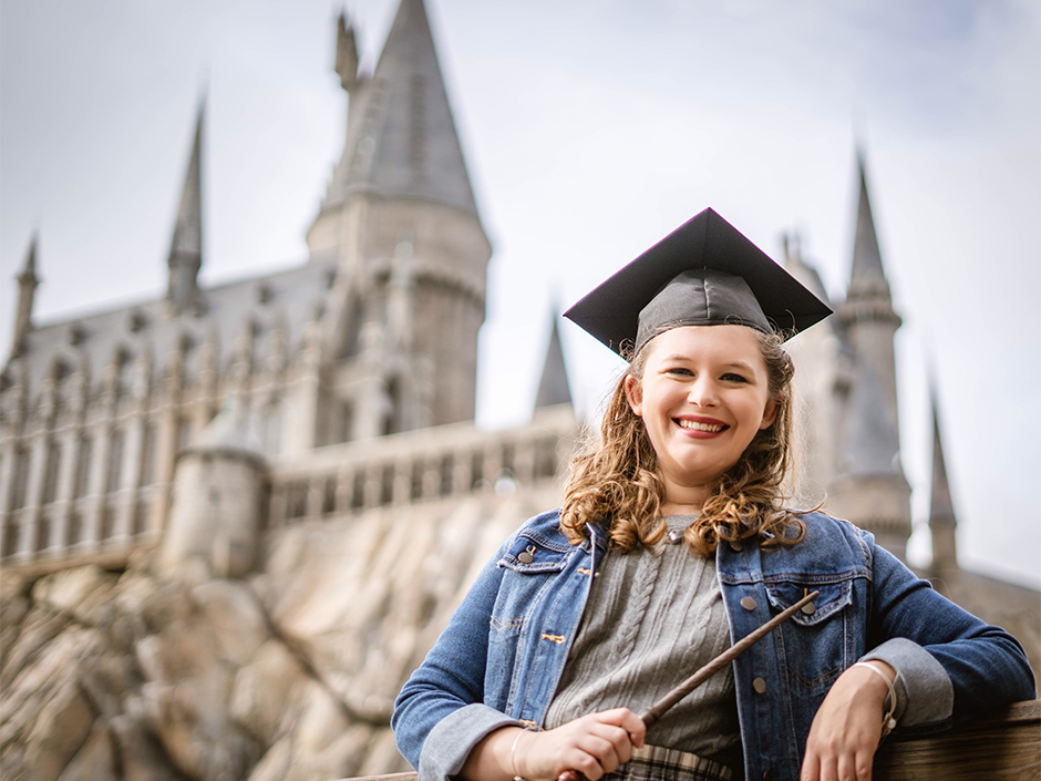 A girl poses with a wand and graduation cap in front of Hogwarts Castle
