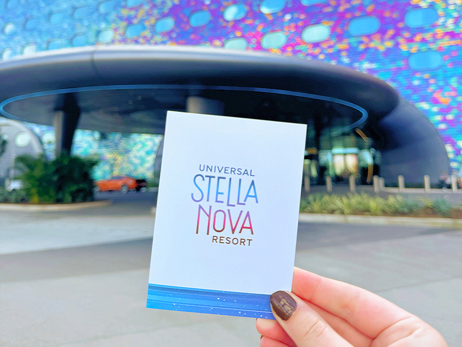 A hand holding up a Stella Nova Resort room key in front of the hotel's entrance
