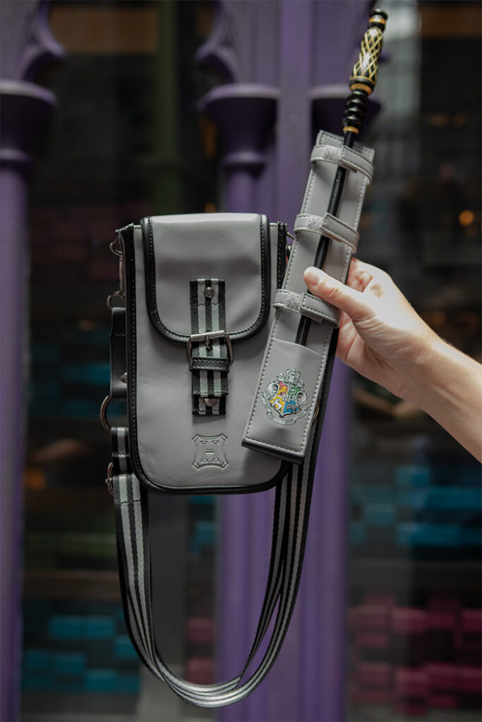 Hand holding up a light gray and black sling bag with a wand holster featuring the Hogwarts crest as the design.