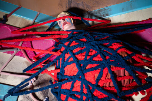 A mannequin doll from Dolls: Let's Play Dead is wrapped tightly in a web of red and blue yarn that crisscrosses over its body and face. The doll has a pale complexion with short blonde hair. Its makeup is dramatic and intentionally smeared with bright blue eyeshadow streaks downward from the eyes like tears. The lips are smeared with vivid red paint. The yarn crosses the face of the doll, covering one eye, adding to the layered, artistic composition. The background features pink heart shapes and teal accents, contributing to the vibrant and surreal atmosphere.