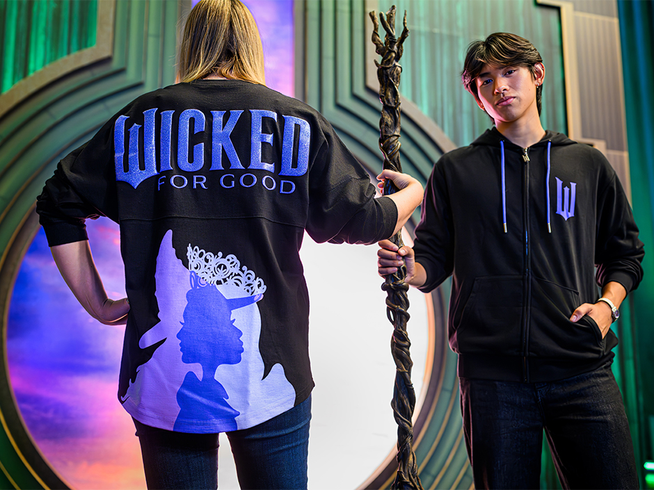 A woman wearing a black and purple "Wicked: For Good" jersey holding a staff next to a man wearing a black and purple "Wicked: For Good" zip-up hoodie inside Wicked: The Experience in Universal Studios Florida.