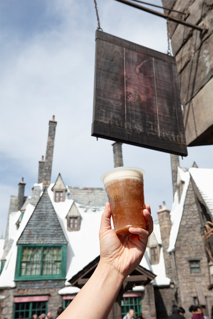 A guest holds up a cup of Butterbeer in front of the Hog's Head sign in The Wizarding World of Harry Potter.