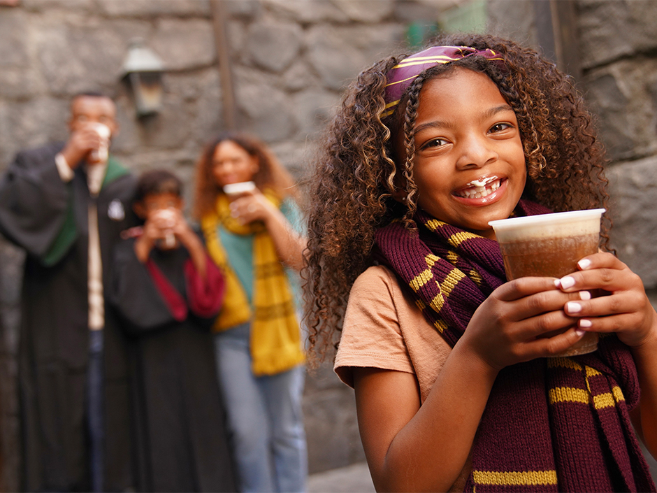 A girl smiles while holding a Butterbeer while her family sips from their own Butterbeer cups in the background