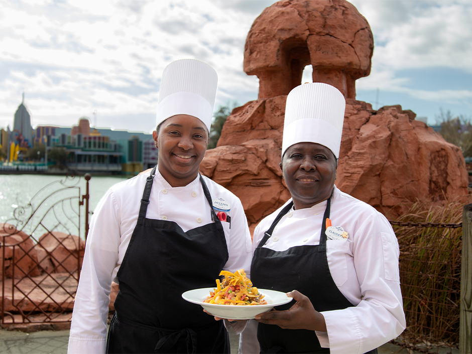 Mythos Restaurant chef assistants Jayann Johnson-Barrett and Claudette Cooper holding a plate of their Women's Herstory Month creation, the Braised Chicken Rasta Pasta.